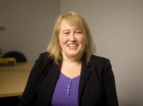 Woman sitting infront of a table with a black blazer and purple blouse